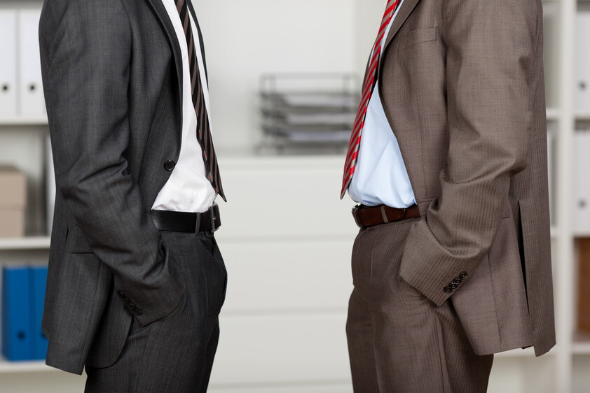 2 Männer in Anzügen und Hände in den Taschen stehen sich gegenüber.