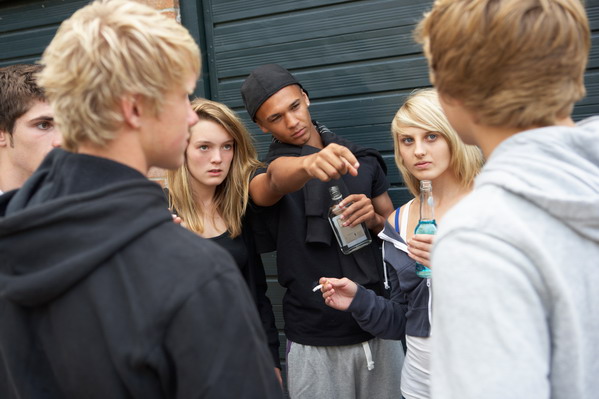 Eine Gruppe Jugendlicher trinkt Alkohol und raucht. Einer von Ihnen zeigt auf einen anderen Jungen und guckt ihn scharf an.